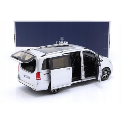 Rear-left three‑quarter view of a 1:18 scale Mercedes‑Benz V‑Class (W447) AMG Line 2018 diecast model in brilliant silver metallic displayed in front of its dark blue Norev box, showing the tailgate lifted and left sliding door open revealing the detailed interior, with black roof rails, panoramic roof, and multi‑spoke wheels visible.